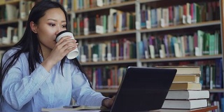 年轻的亚洲学生女孩坐在大学图书馆的一堆书的桌子上，在笔记本电脑上工作，喝咖啡微笑着看着别处，准备考试