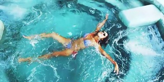 顶部的观点美丽的年轻女子在比基尼和太阳镜放松在游泳池在水晶清澈的水。缓慢的运动。1920 x1080