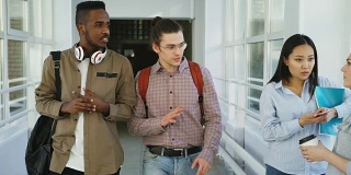 白人嬉皮士和非裔美国学生走在大学宽敞的玻璃走廊里讨论着什么，向女同学挥手，然后继续前进