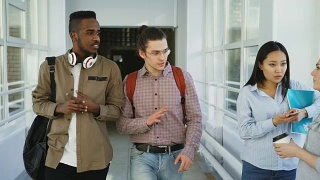 白人嬉皮士和非裔美国学生走在大学宽敞的玻璃走廊里讨论着什么，向女同学挥手，然后继续前进视频素材模板下载