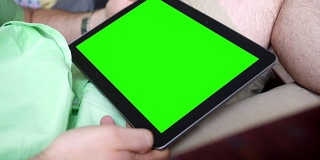 一个白人男性在客厅扶手椅上看一个普通的绿色屏幕平板电脑