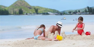 家庭在热带白色沙滩上建造沙堡。父亲和两个女孩在热带海滩上玩沙子