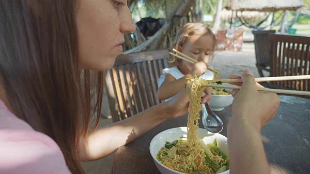 母女在露天的海滩咖啡馆用筷子吃面条汤