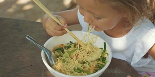 小女孩在露天的海滩咖啡馆用筷子吃面汤