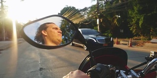 年轻的大胡子男子驾驶摩托车在热带岛屿在美丽的日落慢动作旅行。泰国。方向盘的视图。1920 x1080