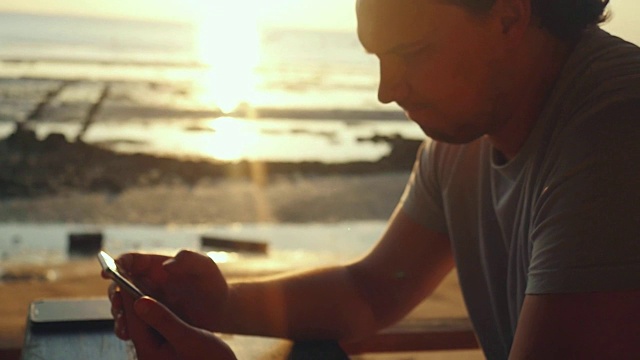 帅哥在海滩上的酒吧休息，在美丽的日落慢镜头中使用手机。1920 x1080