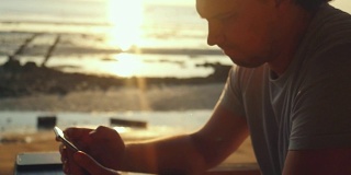 帅哥在海滩上的酒吧休息，在美丽的日落慢镜头中使用手机。1920 x1080