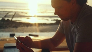 帅哥在海滩上的酒吧休息，在美丽的日落慢镜头中使用手机。1920 x1080视频素材模板下载