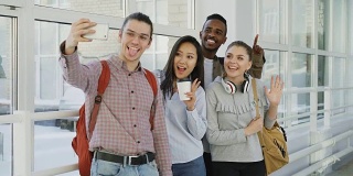 一组四名多民族阳性男女学生正站在宽阔的走廊上。潮人肚子拿着智能手机自拍