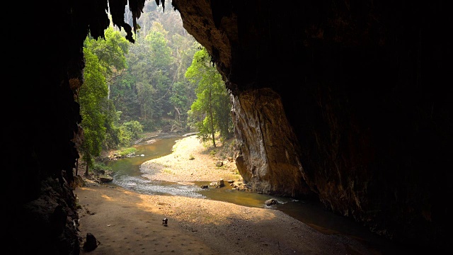 一个老妇人在一个大洞的底部收集蝙蝠粪，洞里有一条河。泰国Tham Lod Cave, Pai。