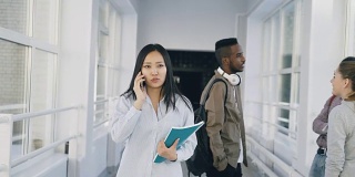 照片中，亚洲女学生走在宽阔的大学走廊上，一边打电话一边与其他多民族学生擦肩而过
