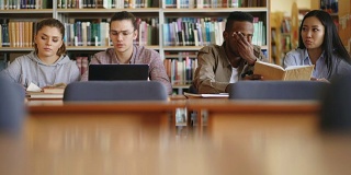 多民族学生坐在图书馆里，桌上放着书和笔记本电脑，一起笑着准备考试