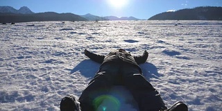 年轻人躺在雪地上，在阳光明媚的日子里做天使。快乐的人在白雪覆盖的草地上嬉戏微笑。男孩享受自由，享受旅行的乐趣。背景是阳光。特写慢动作