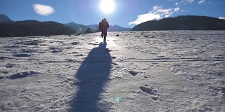 在阳光明媚的冬日，一个面目全非的家伙在雪地上慢跑。年轻人在白雪覆盖的草地上奔跑。背景是阳光和蓝天。假日或假期的概念。后视图慢动作