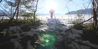 在森林附近白雪覆盖的草地上行走的难以辨认的徒步旅行者。一个年轻人在阳光明媚的冬日在雪地上徒步旅行。背景是阳光。旅行或旅行的概念。后视图慢动作