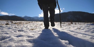 在阳光明媚的日子里，一个不认识的人用棍子爬上草地的雪山的后视图。年轻的徒步旅行者在雪地上徒步旅行，享受冬季旅行。健康积极的生活方式。慢动作