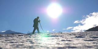 年轻的徒步旅行者走北欧与棍棒在雪地上在太阳的背景。一个不认识的人在雪地上跋涉，在阳光明媚的日子里爬上小山。健康积极的生活方式。侧视图慢镜头
