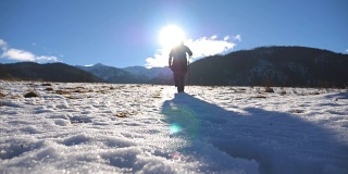 后视图的年轻的徒步旅行者与棍棒爬上雪山在田野。一个面目全非的人走在雪地上享受冬季旅行。阳光在背景。健康积极的生活方式