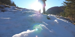 在阳光明媚的日子里，年轻的徒步者爬上雪坡的后视图。冬天，一个面目全非的人走在雪山森林里。健康积极的生活方式。旅行或旅行的概念。慢动作