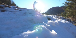 在冬季的旅途中，一个不知名的游客在雪坡上攀爬。在阳光明媚的日子里，年轻的徒步旅行者在山林中行走。背景是阳光。旅行的概念。后视图慢动作