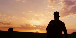 一个少年背着背包走向田野或乡村的夕阳。剪影视频，后视图。概念——新的研究，向未知的方向前进，离开家