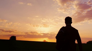 一个少年背着背包走向田野或乡村的夕阳。剪影视频，后视图。概念——新的研究，向未知的方向前进，离开家视频素材模板下载