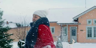 快乐的年轻微笑的女人扔雪球，并被一个击中。穿红外套的女孩和她的伴侣享受冬天。在缓慢的运动。田园小屋后院里的一天。