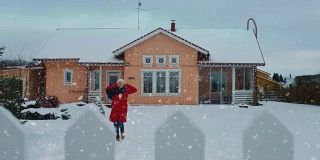 美丽的年轻微笑女人跳跃和旋转下飘落的雪。穿红外套戴围巾的女孩在后院享受冬天。田园诗般的房子站在背景。