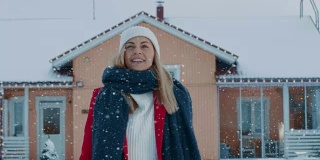 一个年轻的金发女人的肖像在红色大衣与围巾在她的后院行走和跳跃，而雪花落。背景是田园诗般的房子，带有冬季景色。
