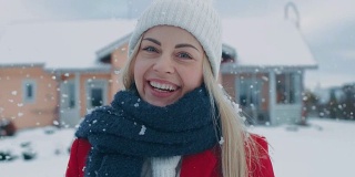 一个年轻的金发女人的肖像在红色大衣与围巾微笑进入相机，而站在飘落的雪。在后院田园诗般的房子与冬天的景色。