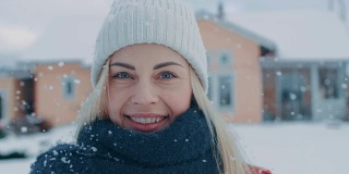 一个年轻的金发女人的肖像在红色大衣与围巾微笑进入相机，而站在飘落的雪。在后院田园诗般的房子与冬天的景色。