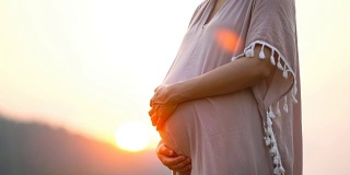 怀孕亚洲女性触摸她的腹部在粉红色的裙子与日落的背景