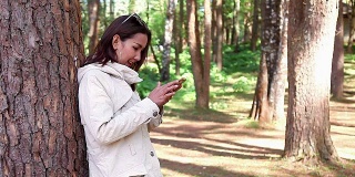 年轻美丽的女人Lean the Trees和她的手机聊天穿长外套夹克在寒冷的天气在国家松树森林公园与幸福。