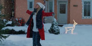美丽的年轻微笑的女人在飘落的雪纺纱。在她田园诗般的房子的前院，穿着红外套、围着围巾的女孩正在享受冬天。