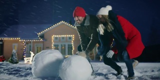 快乐的年轻夫妇堆雪人，都滚大雪球。一家人在一个冬天的晚上玩耍。在装饰着花环的背景房子里。
