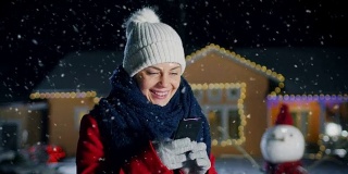 平安夜，美丽的年轻女子站在用花环装饰的田园别墅的后院使用智能手机。戴围巾穿红外套的女孩，用手机给家人和朋友发圣诞短信。