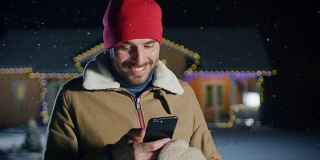 平安夜，一名英俊的年轻人站在自家用花环装饰的田园别墅的后院使用智能手机。一个家伙用手机给他的家人和朋友发圣诞短信。