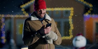 平安夜，一名英俊的年轻人站在自家用花环装饰的田园别墅的后院使用智能手机。一个家伙用手机给他的家人和朋友发圣诞短信。