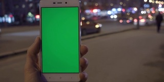 男子手握白色智能手机，在夜城灯光散景的背景下。绿色的屏幕。