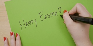 女人用红色的指甲在绿色的纸上用黑色的记号笔写着复活节快乐的大写字母