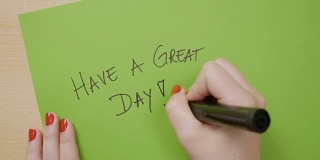 一名年轻女子，手指上涂着红色指甲油，用黑色记号笔在绿纸上写下美好的一天