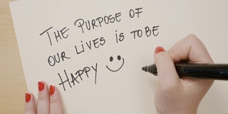 女性的手写我们生活的目的是幸福励志的引用和画一个笑脸在白纸上用黑色马克笔