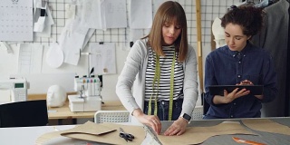 一位女服装设计师正在用粉笔在布料上画出新衣服的轮廓，而她的同事正在用写字板帮助她。技术在服装制造的概念。