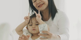 泰国可爱的小女孩和她的妈妈在卧室用智能手机自拍的现代生活方式