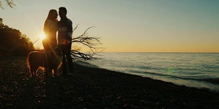 年轻夫妇在日落与宠物。肩并肩站在湖的背景和夕阳