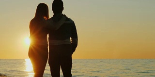 一对年轻的夫妇在海边拥抱着欣赏夕阳的剪影。后视图
