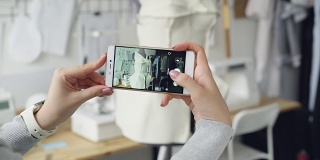 近距离拍摄的女人的手拿着智能手机，拍摄裁缝假人与半成品衣服别在上面。现代技术在小型企业的概念。