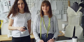 两个女服装设计师企业家站在一起，看着相机的肖像。一个女人拿着外卖咖啡，另一个摸着演播室的桌子。