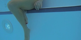 水下摄影机接近坐在游泳池台阶上的女孩的下半身