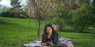 微笑的年轻女子躺在草坪上，手里拿着平板电脑